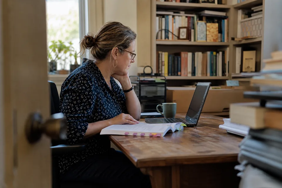Community college instructor at a desk reviewing a printed course reader next to a laptop
