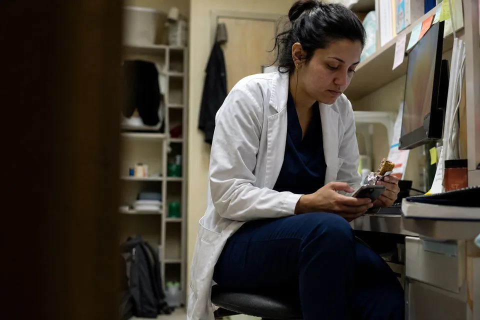 Physician assistant trainee in a short white coat answering practice questions on a phone in a cluttered hospital workroom