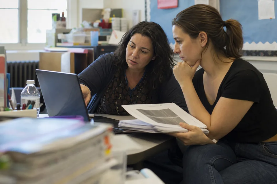 Two teachers in a staff workroom planning a review game from printed handouts