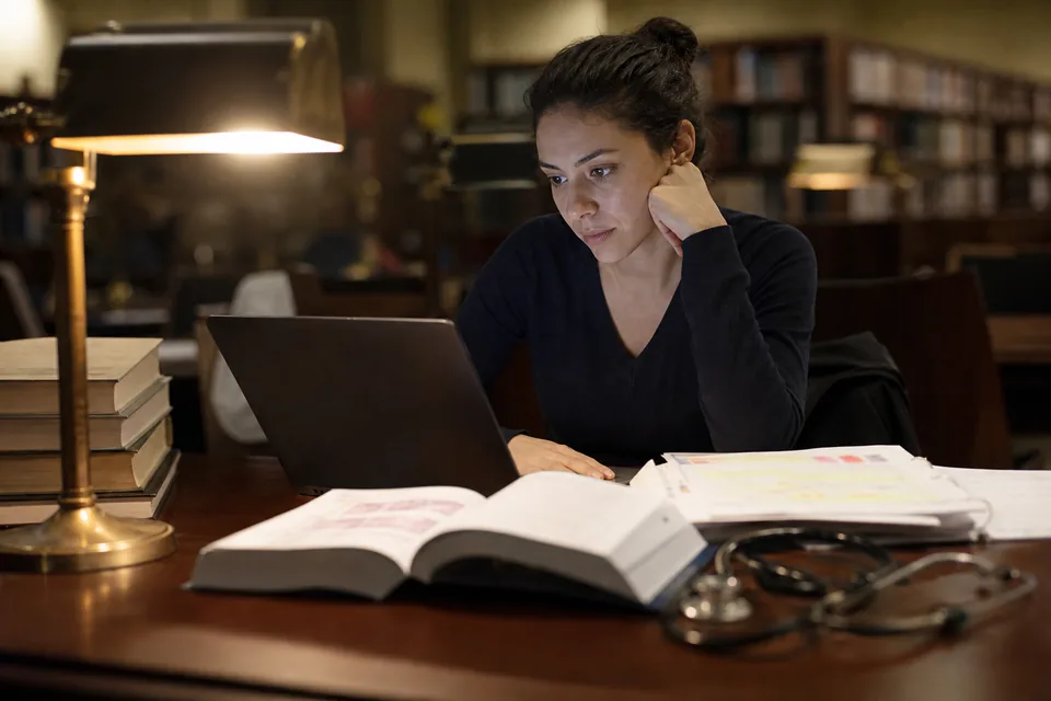 Future physician reviewing lecture slides and a pathology review book at a library desk in the evening